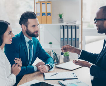 Side view portrait of couple is embracing, getting a key from their future apartment from a broker, and signing contract, all are dressed in formal outfits, sitting in workplace, workstation